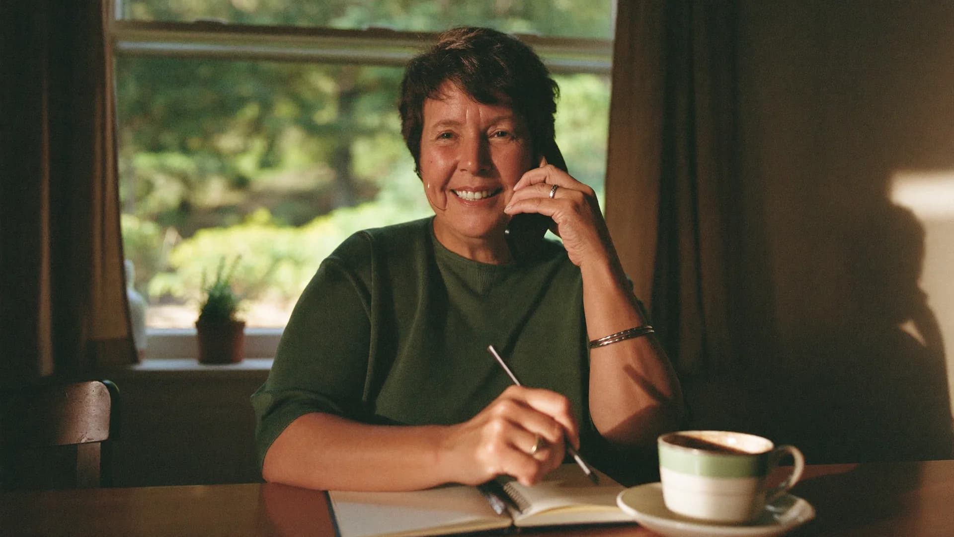 A woman speaks warmly on the phone at a kitchen table with a notepad in front of her